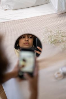 A young woman takes a selfie, capturing her reflection in a tabletop mirror indoors.