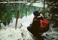 一个背着红色背包的人蹲在冰封的湖边，四周是被白雪覆盖的树木