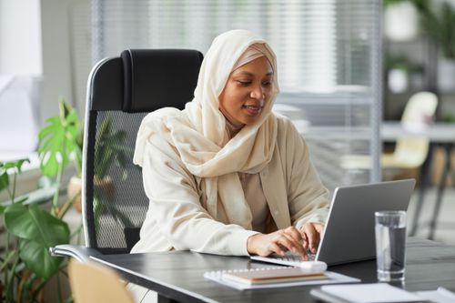 Free Professional Muslim businesswoman in hijab working on laptop in a modern office setting. Stock Photo