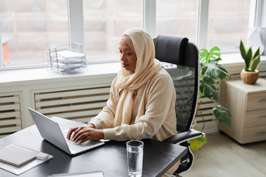 A focused Muslim woman in hijab working on a laptop in a modern office setting. Professional and dedicated.