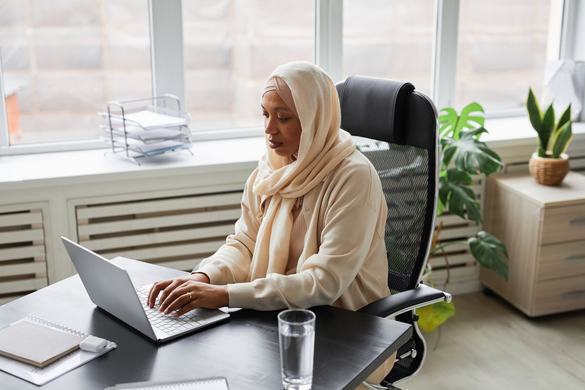 A focused Muslim woman in hijab working on a laptop in a modern office setting. Professional and dedicated.