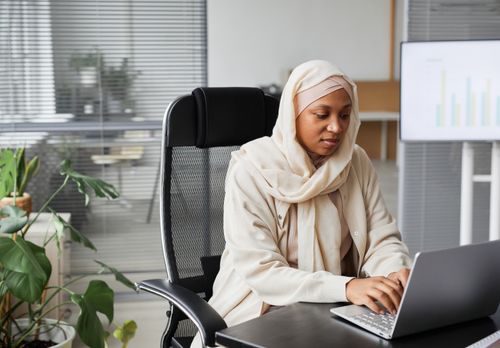 Free Professional Muslim woman in hijab working on laptop in modern office setting, demonstrating focus and dedication. Stock Photo