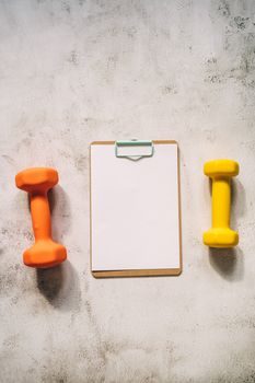 Vertical shot of orange and yellow dumbbells by a blank clipboard on concrete surface.