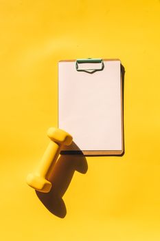 A minimalistic arrangement of a clipboard and dumbbell on a bright yellow background.