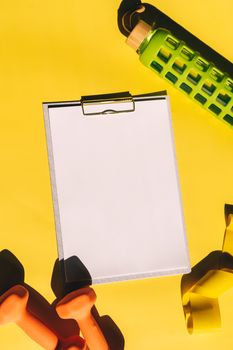 A vibrant fitness flat lay with blank clipboard, dumbbells, and water bottle over yellow background.