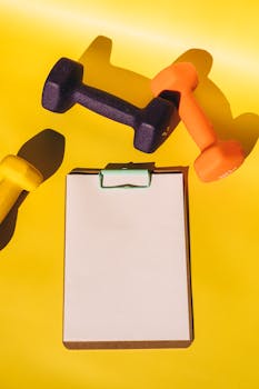 Vibrant still life of multicolored dumbbells and a blank clipboard on a yellow backdrop.