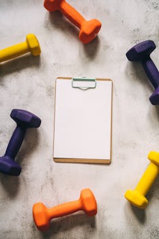 Top view of colorful dumbbells encircling a blank clipboard, offering copy space for text.