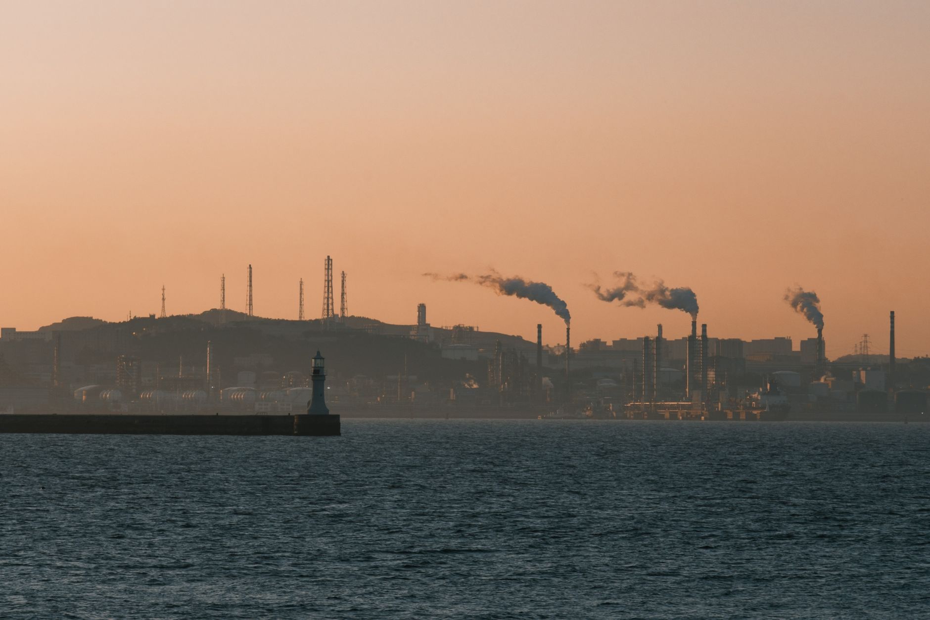 Factory skyline emitting smoke over the ocean at sunset, under an orange sky.