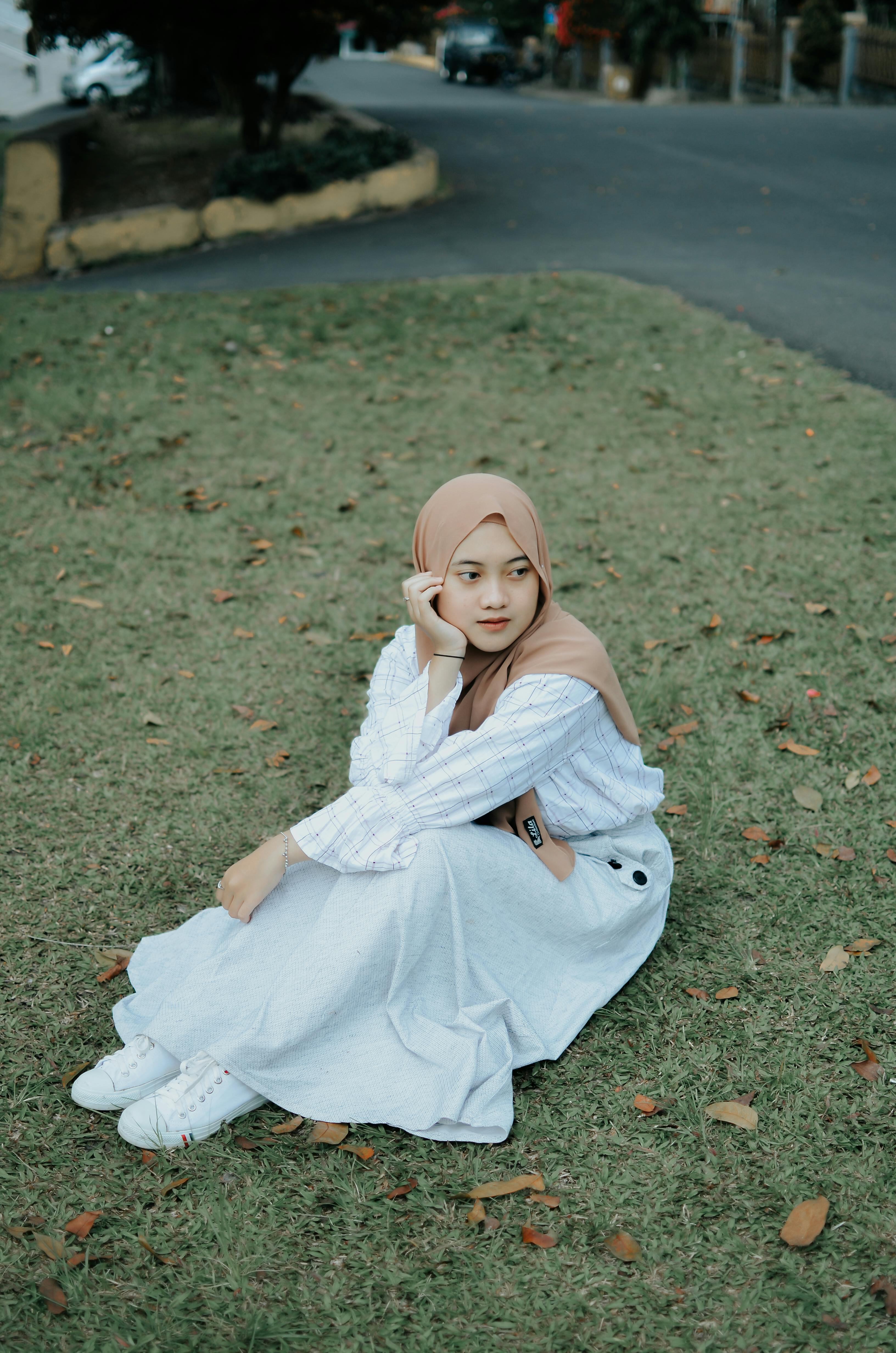 A young Muslim woman wearing a hijab and white dress is sitting on the grass outdoors, creating a serene and peaceful scene.