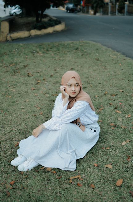 A young Muslim woman wearing a hijab and white dress is sitting on the grass outdoors, creating a serene and peaceful scene.