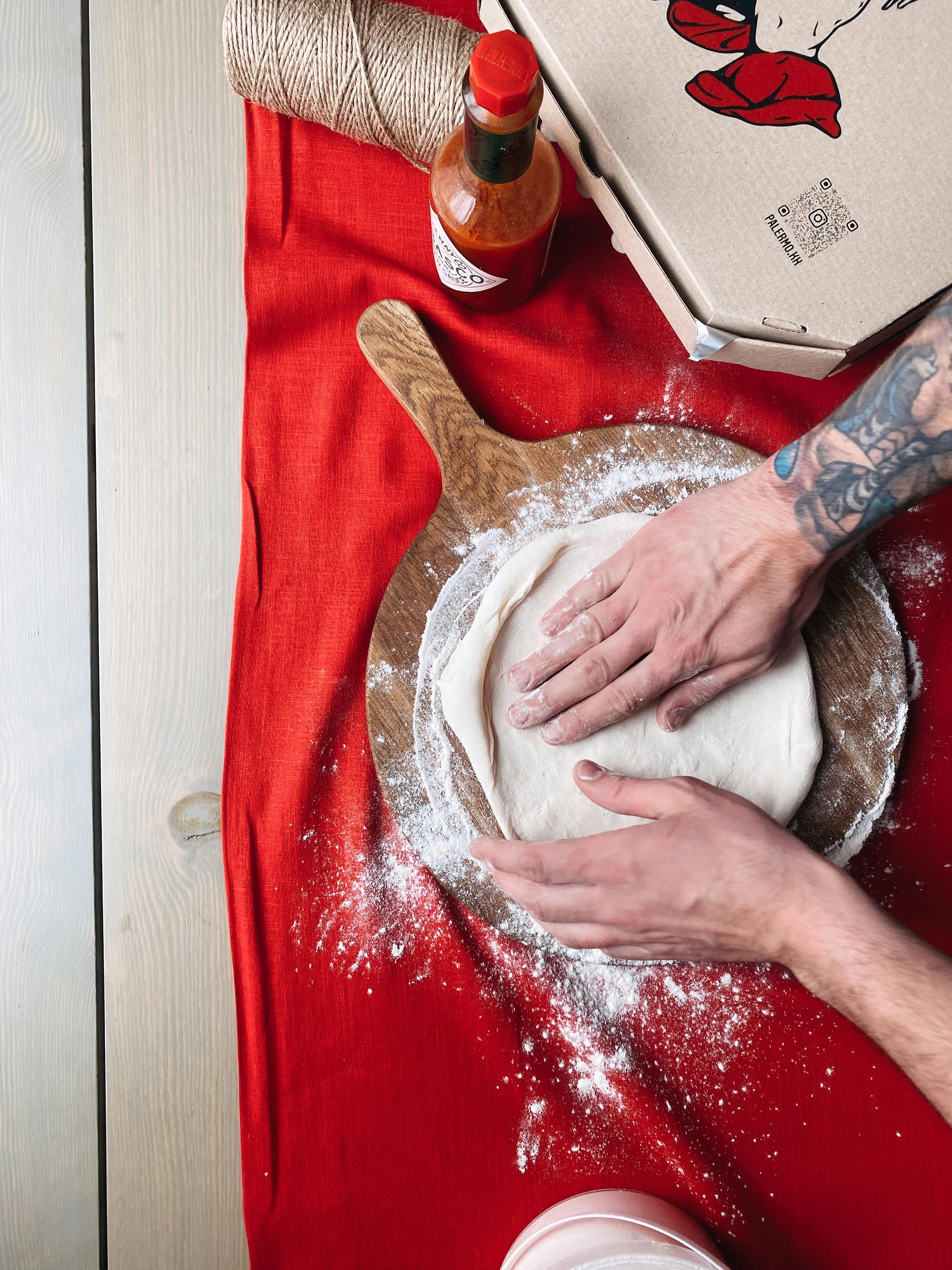 Hands kneading pizza dough on a wooden board with flour and hot sauce nearby.
