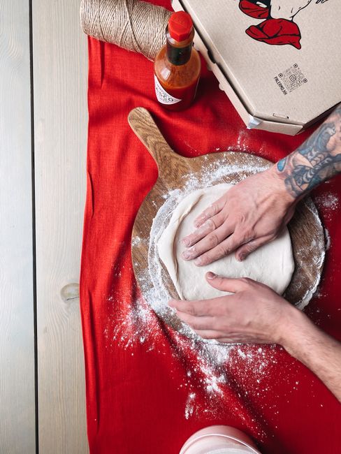 Hands kneading pizza dough on a wooden board with flour and hot sauce nearby.