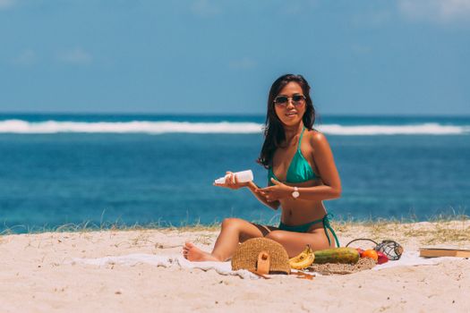 Asian woman enjoying a beach picnic in summer, wearing a green bikini and sunglasses.