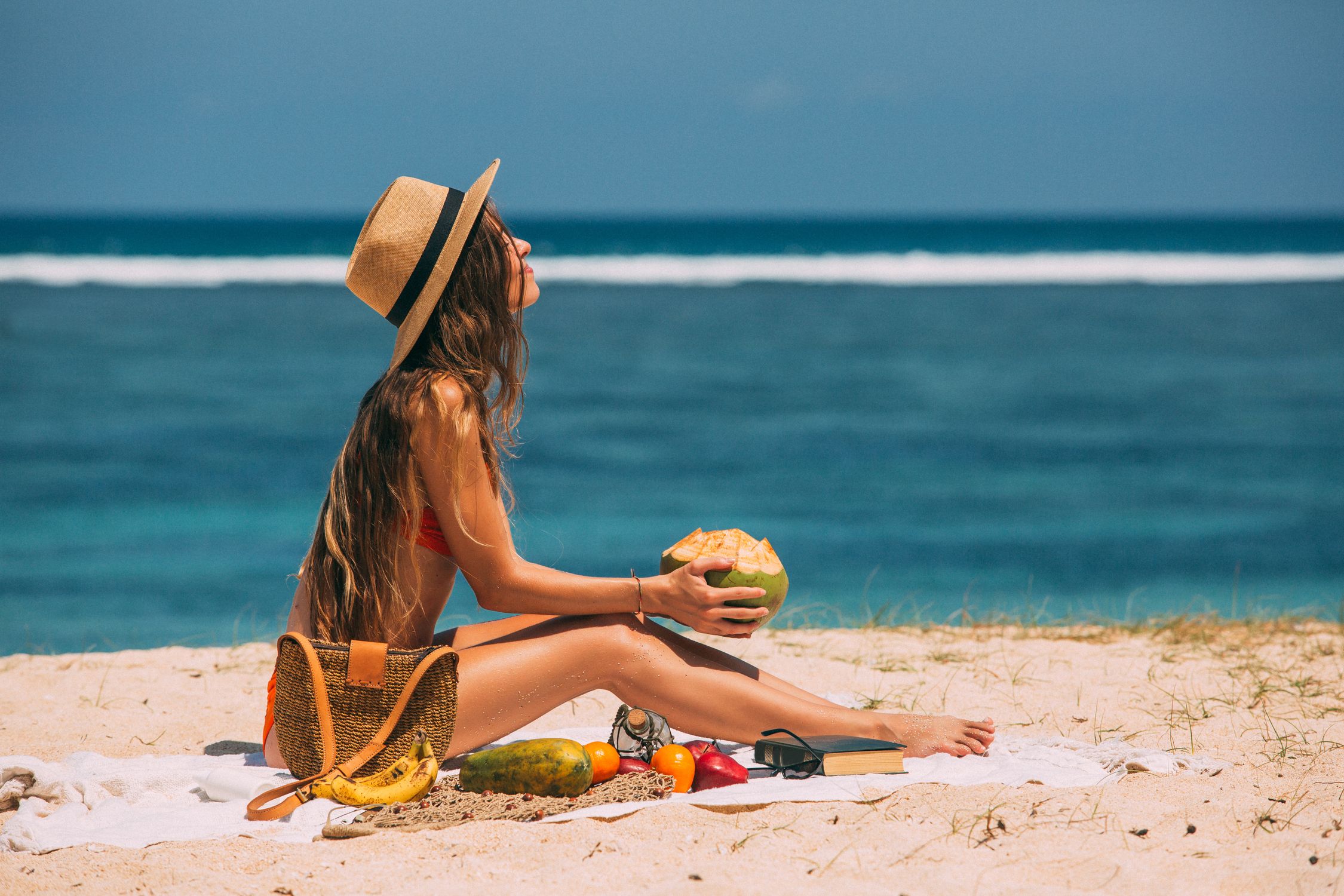 A woman wearing a sun hat holding a coconut while sitting on the sand beside the beach