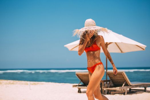 Woman in a red bikini walks on a sunny beach, wearing a straw hat with umbrellas and lounge chairs in the background.