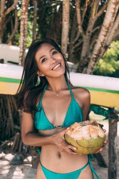 A smiling Asian woman in a blue bikini holds a coconut on a sunny tropical beach.