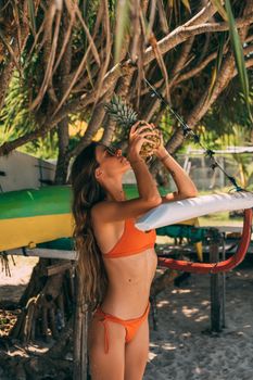 A woman in orange swimwear holding a pineapple under tropical trees at the beach.
