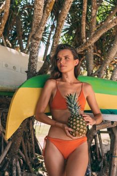 Young woman in orange bikini holding a pineapple under tropical trees.