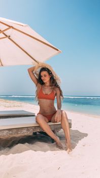 Woman in a red bikini relaxing under a beach umbrella on a sunny day by the ocean.