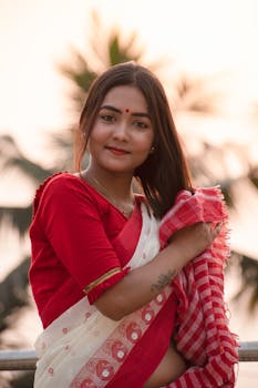 A South Asian woman in a red saree poses outdoors at sunset, embracing cultural attire.
