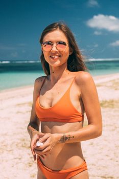 A young woman in an orange bikini and sunglasses standing happily at the beach on a sunny day.