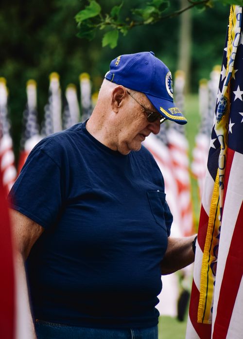 Free A senior veteran in a blue cap stands beside American flags, honoring service. Stock Photo