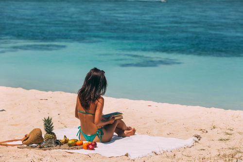Free A woman in a bikini reading a book on a tropical beach, surrounded by fresh fruits. Stock Photo