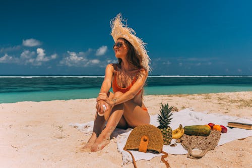 Woman in Orange Bikini with Sun Hat Sitting on Beach Shore