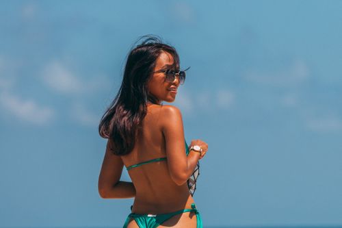 Free An adult woman enjoys a sunny day at the beach, wearing a bikini and sunglasses, looking back with a smile. Stock Photo
