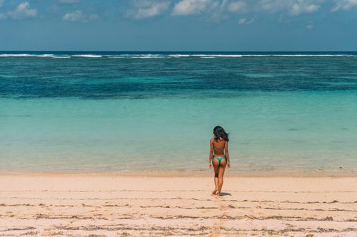Free A woman in a bikini walks towards the turquoise ocean under a sunny sky on a sandy beach. Stock Photo