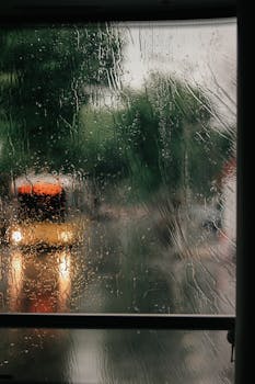 Raindrops on a window with blurred cityscape and vehicle lights. Moody atmosphere.