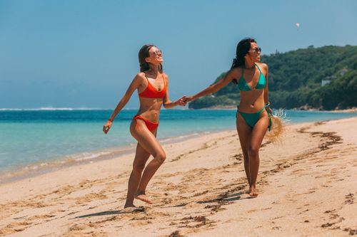 Free Two women in bikinis walking joyfully along a tropical beach, holding hands under the sun. Stock Photo