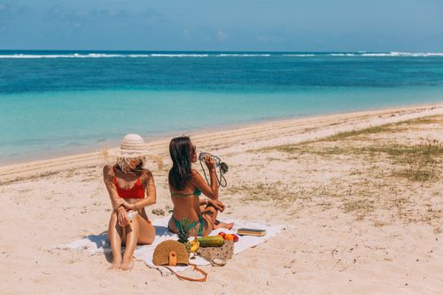 Free Two women enjoy a sunny day at a tropical beach, relaxing with fruits and drinks. Stock Photo