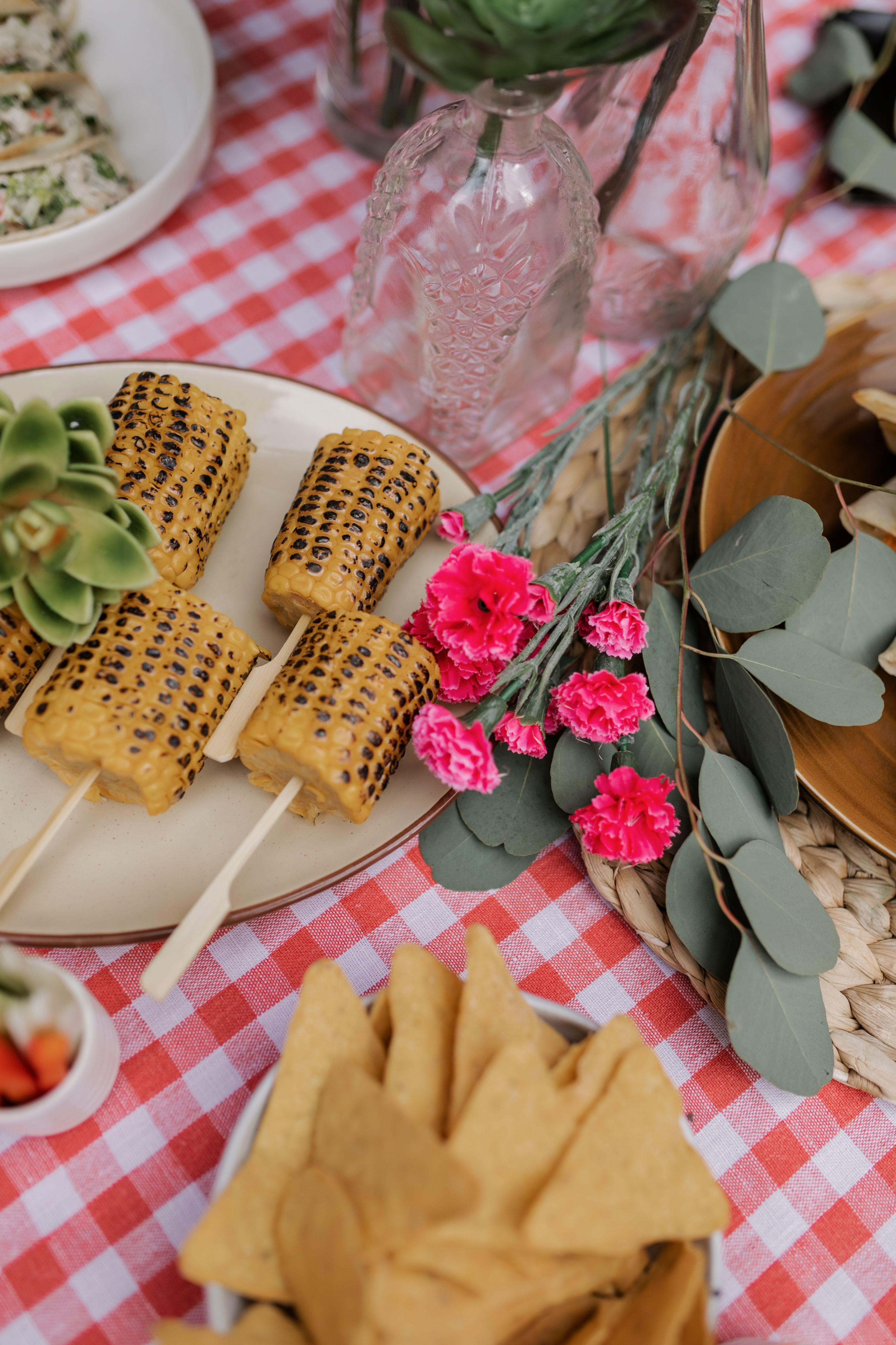 Colorful picnic setup with grilled corn, nachos, and vibrant flowers on a checkered tablecloth.