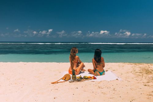 Free Two women in bikinis relaxing on a sandy beach with a tropical fruit picnic under a clear blue sky. Stock Photo