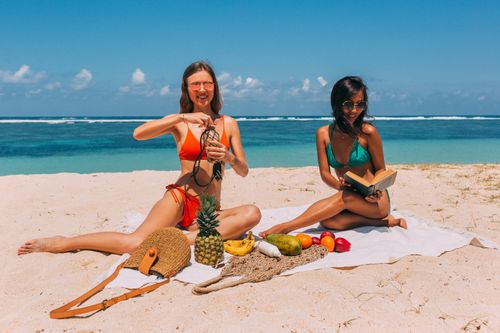 Free Two women in vibrant swimsuits enjoy a sunny beach picnic with fresh fruit and books. Stock Photo