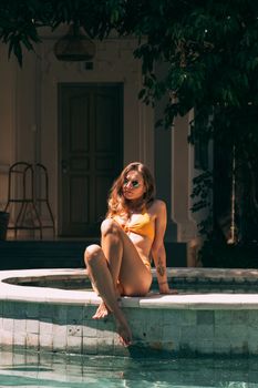 Woman in swimwear relaxing poolside, basking in sun, wearing sunglasses and enjoying leisure time.