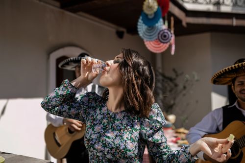 Free Woman in floral dress drinking tequila outdoors with musicians at a festive gathering. Stock Photo