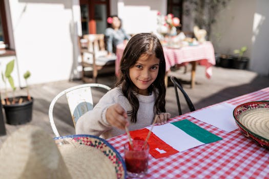 A smiling girl paints a Mexican flag at an outdoor table, enjoying a sunny day.