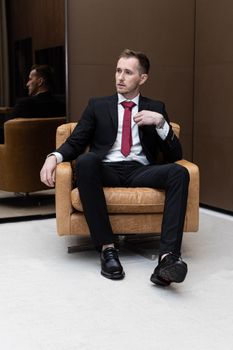 Portrait of a man in a black suit and red tie sitting on a modern chair indoors.