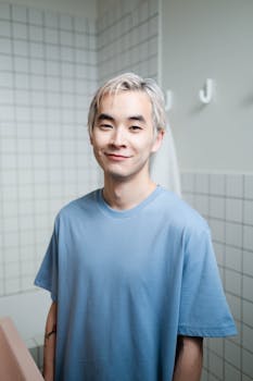 Portrait of a smiling young man in a casual blue t-shirt standing in a bathroom with tiled walls.