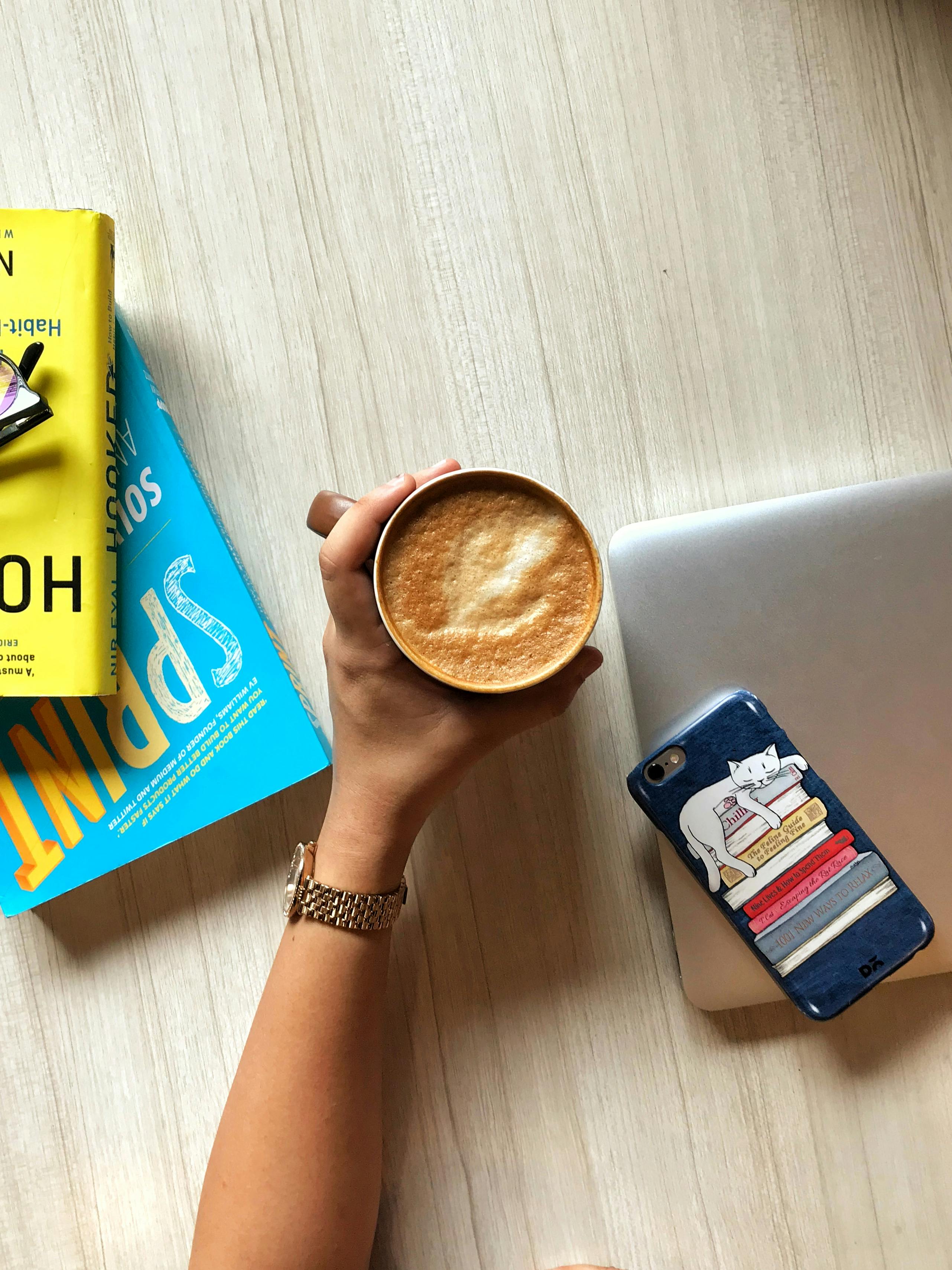 从顶视图看，一只手持拿铁咖啡放在木桌上，旁边有书籍和手机