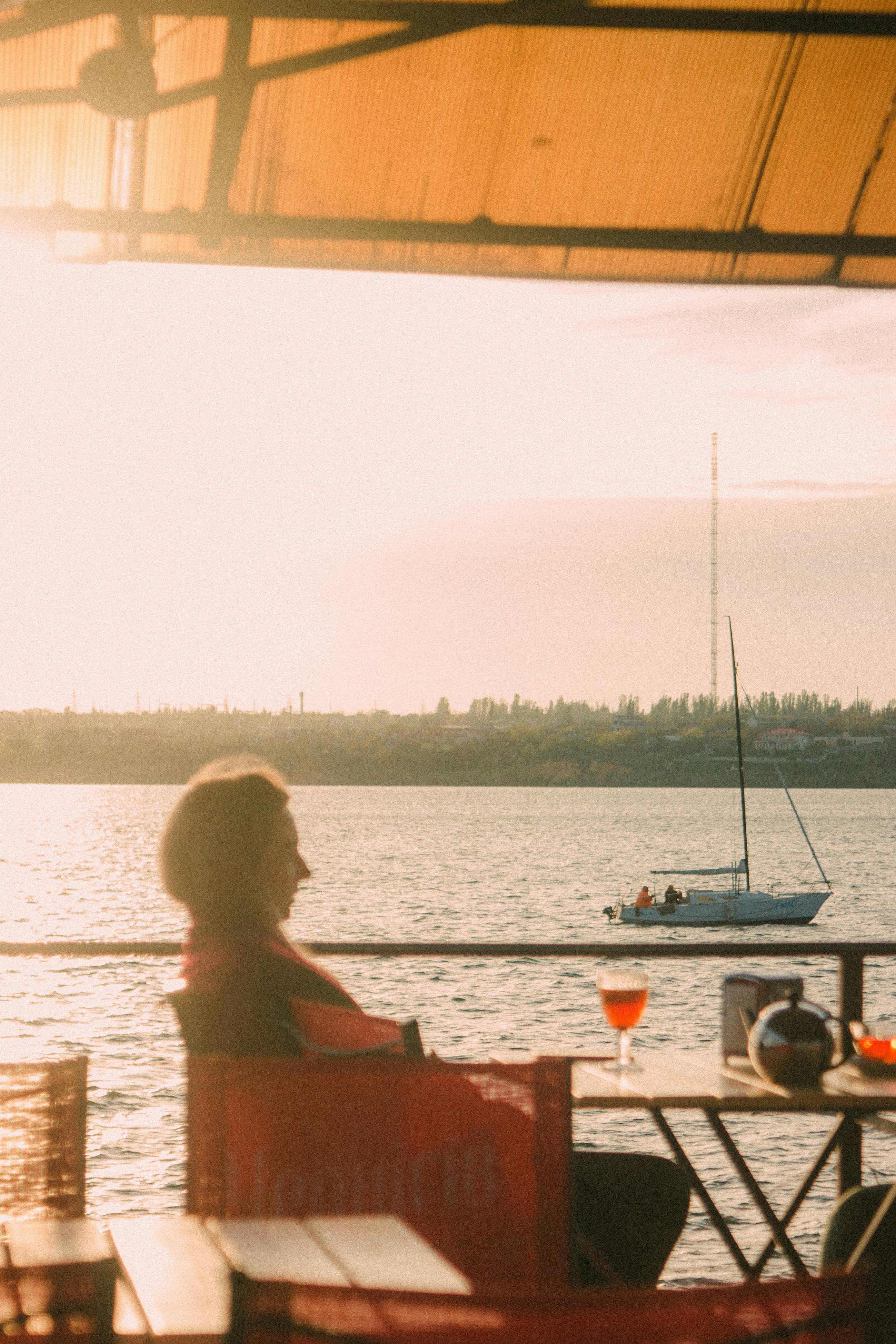 Side view of female sitting on terrace at table with wineglass of alcohol drink in outdoor cafe on waterfront near river with floating boat
