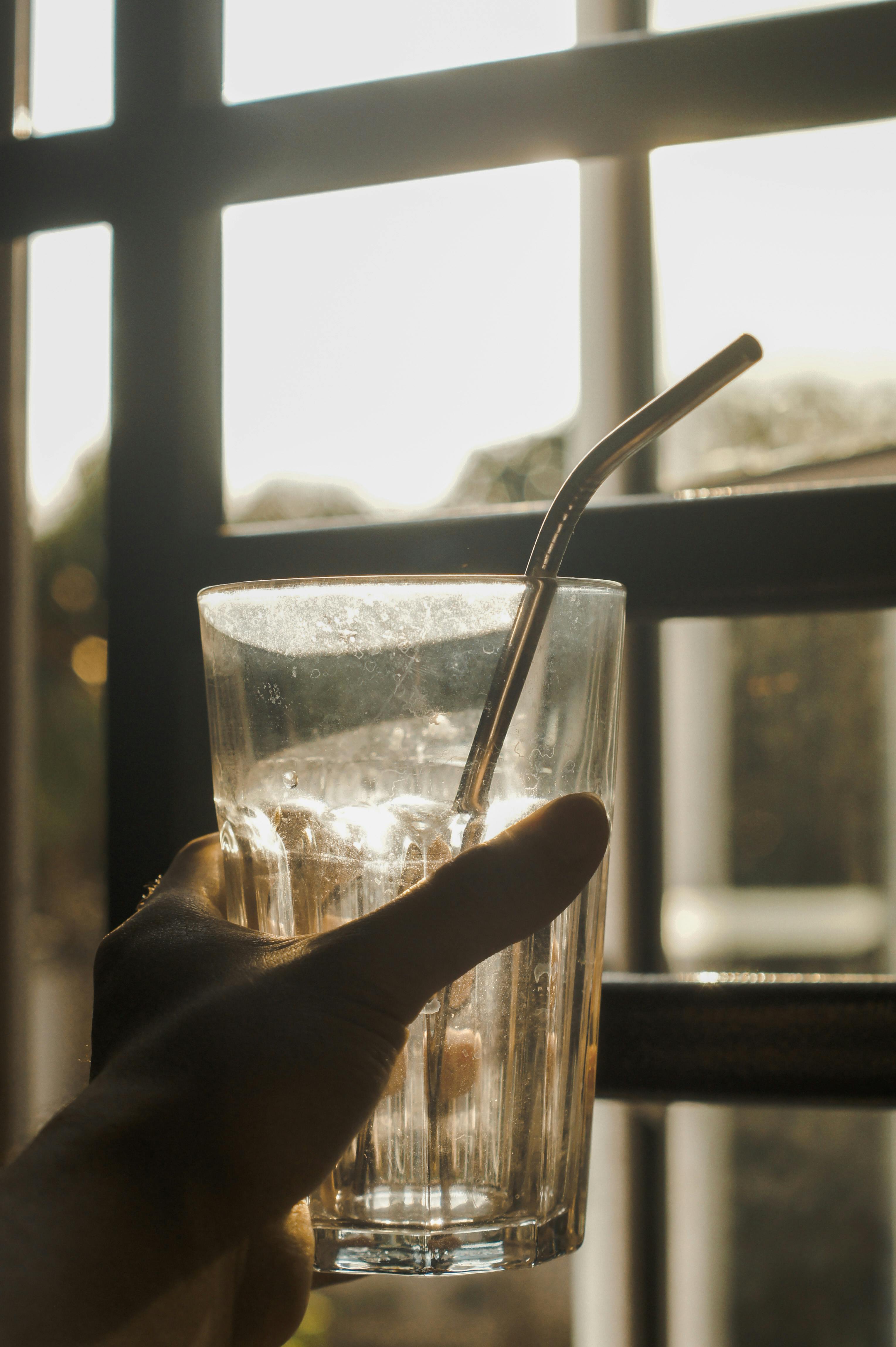 Close-up of a hand holding a glass with a metal straw by a sunlit window, perfect for sustainability themes.