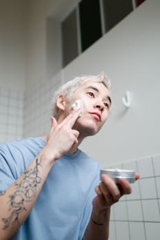 Asian man with tattoo applying skincare cream as part of morning routine in bathroom.