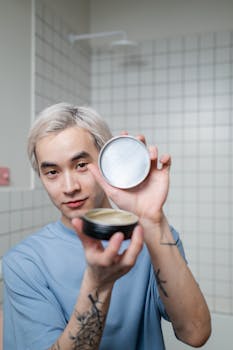 A young man with blonde hair shows off a hairstyling product in a modern bathroom.