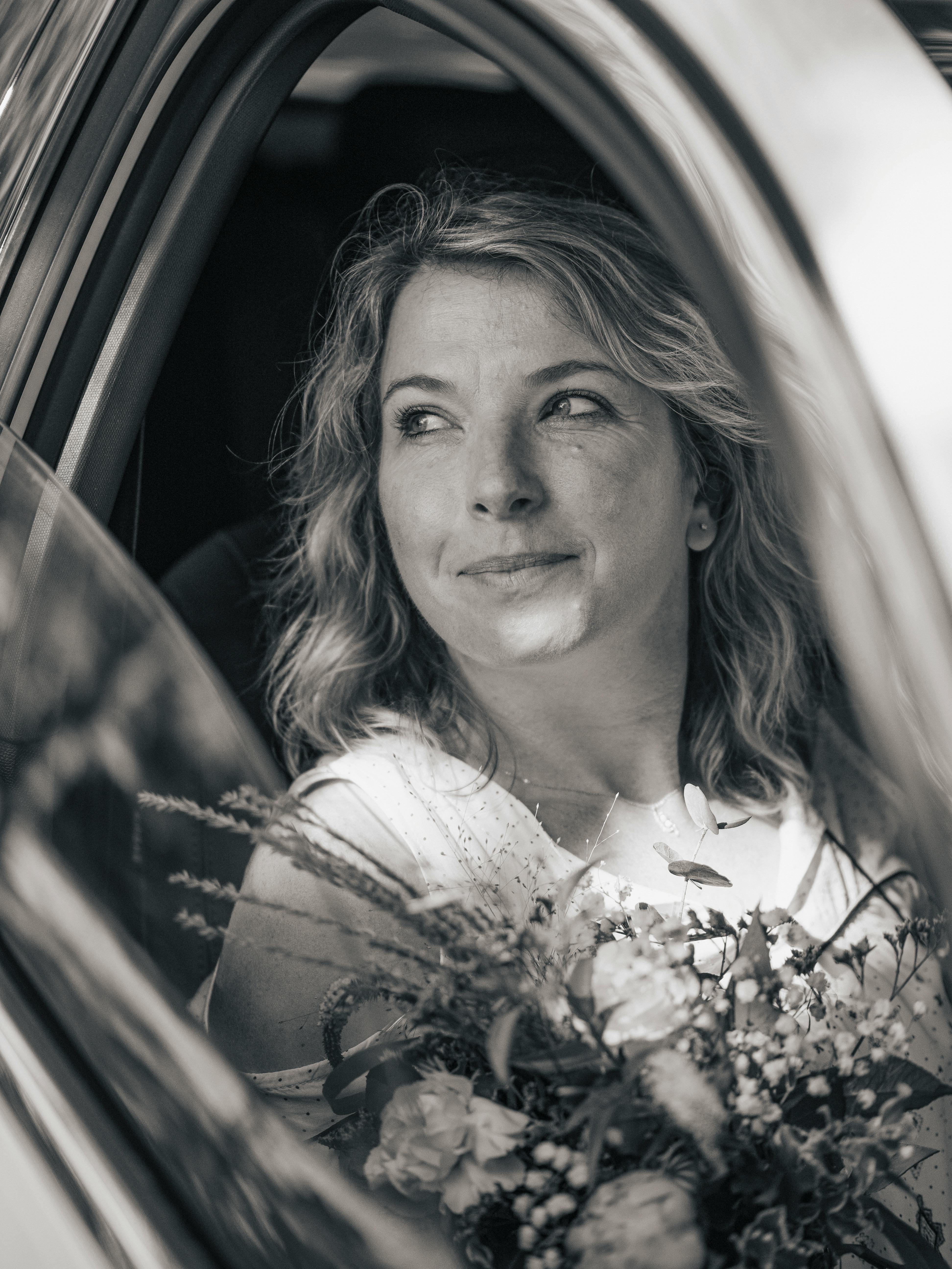 Black and white portrait of a smiling woman holding flowers through a car window.