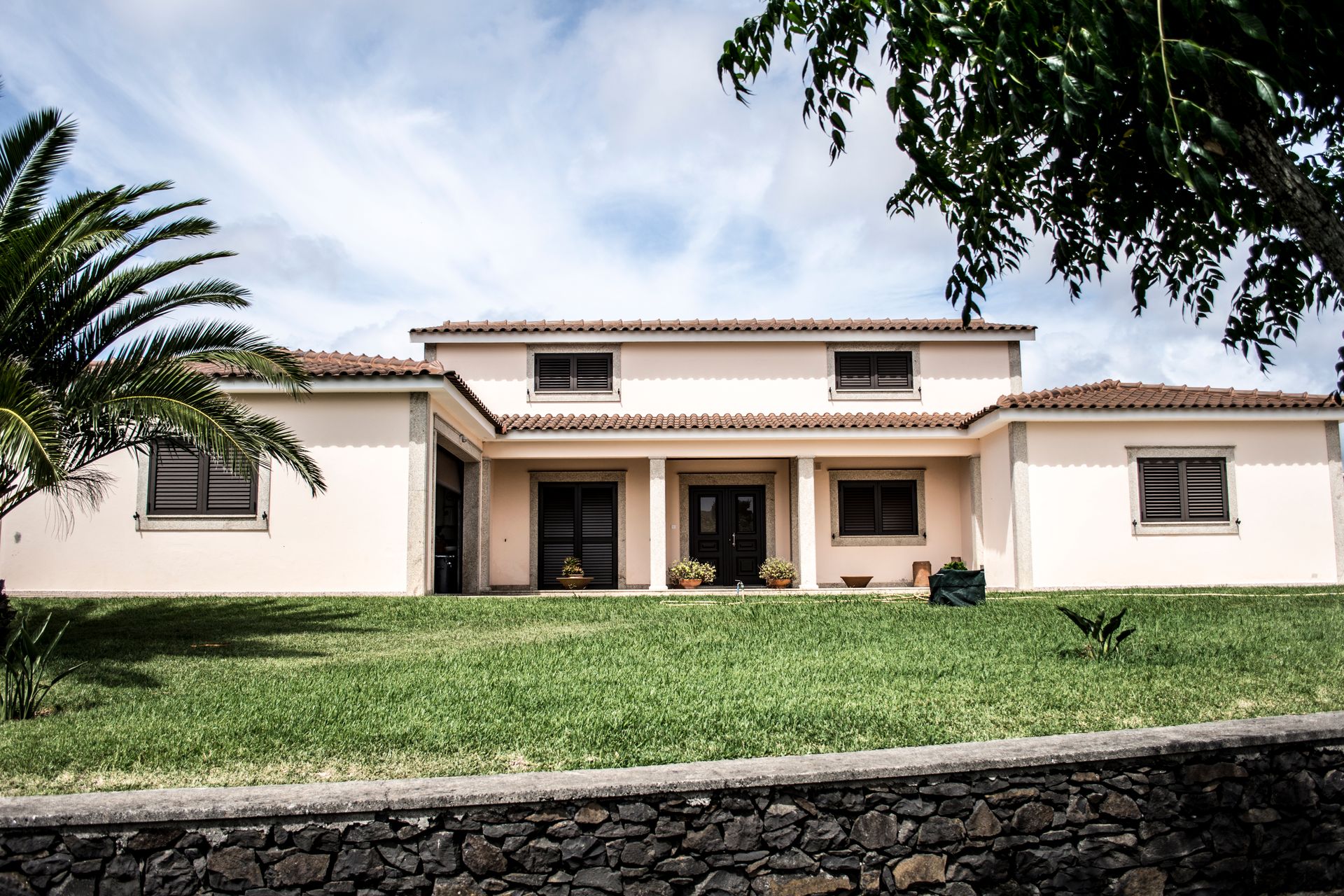Stucco home with a tile roof and a tall palm tree behind a low stone wall