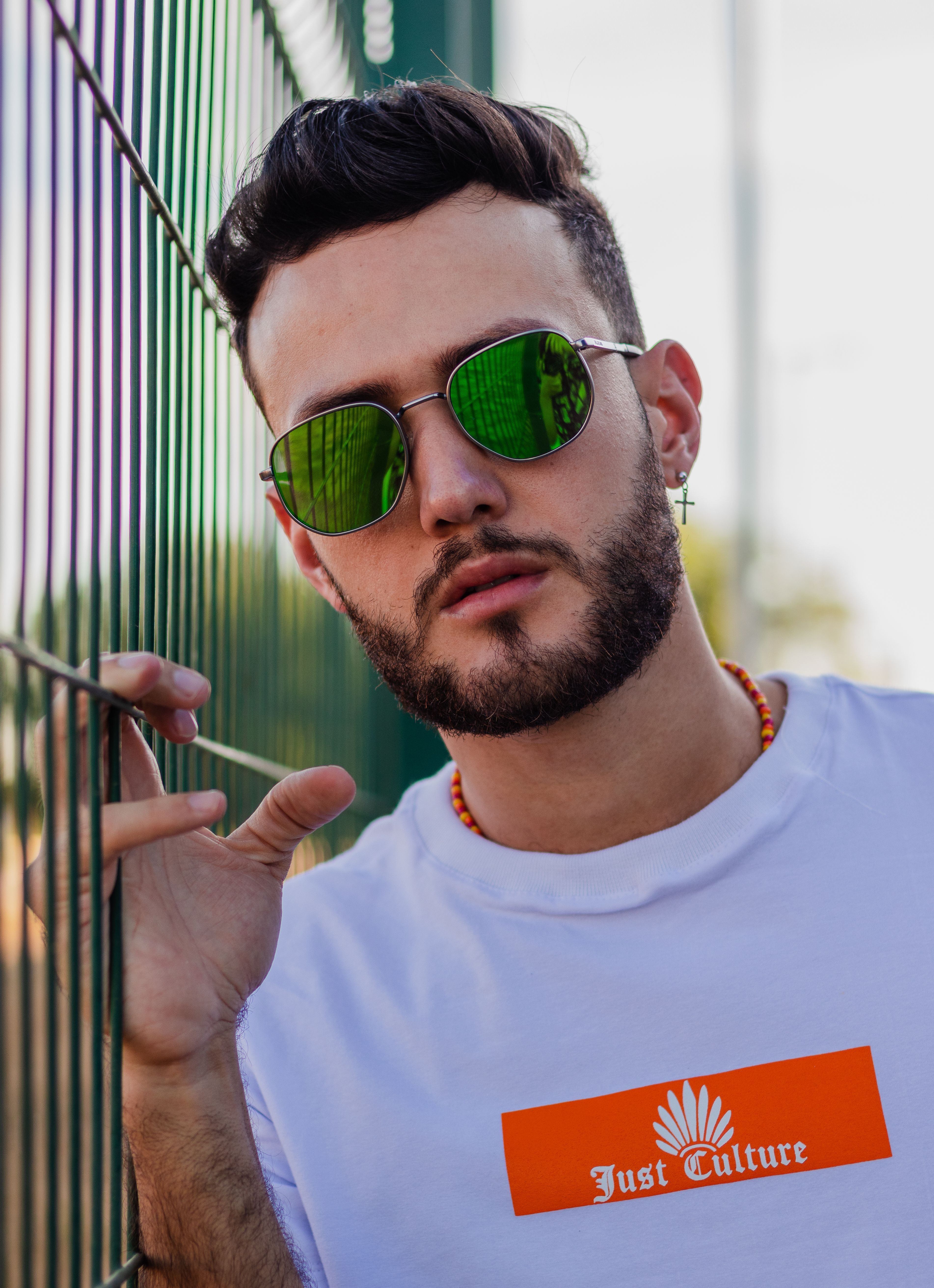 Stylish man with sunglasses and beard poses by a fence in casual wear.