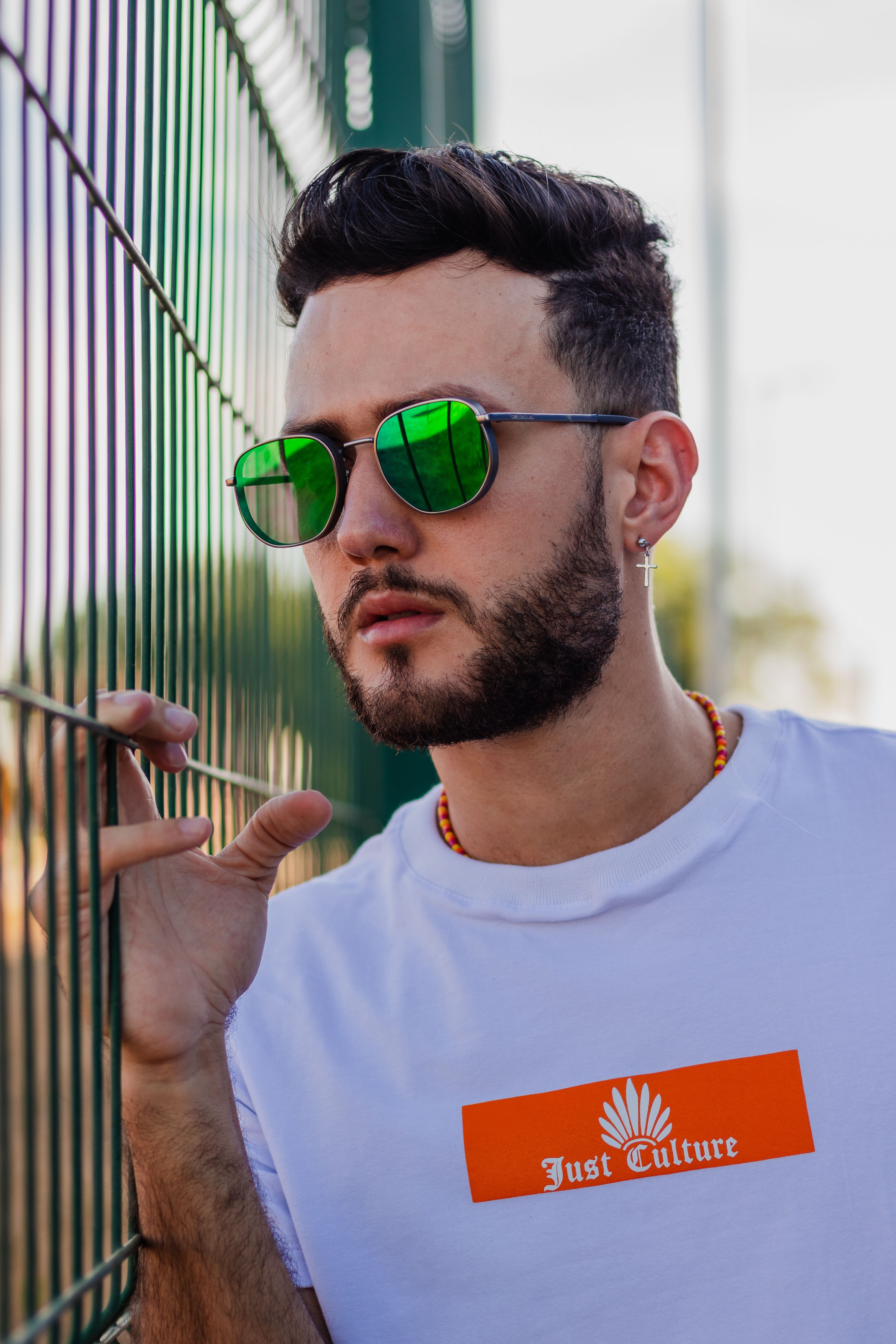 Modern portrait of a bearded young man with green sunglasses by a metal fence.